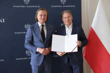 Dwóch mężczyzn w garniturach stoi na tle granatowej ściany z napisem „WOJEWODA MAZOWIECKI”. Jeden z nich trzyma otwartą teczkę z dokumentem. Obok nich widoczna jest flaga Polski.