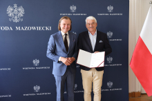 Dwóch mężczyzn stoi na tle granatowej ściany z napisem „WOJEWODA MAZOWIECKI” i godłem Polski. Jeden z nich trzyma otwartą teczkę z dokumentem. Po prawej stronie widoczna jest flaga Polski.