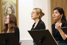 Trzy kobiety w czarnych bluzkach stoją obok siebie z pulpitami na nuty. Kobieta po lewej ma długie, falowane włosy. Środkowa ma blond kok i kolczyki w kształcie gwiazdek. Prawa również ma takie kolczyki i trzyma rękę przy twarzy. W tle drewniane panele i biała ściana z panelem elektrycznym.