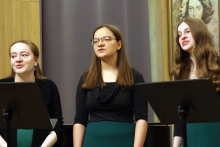 Trzy kobiety w czarnych bluzkach i zielonych spódnicach stoją obok siebie przed pulpitami na nuty. Lewa ma spięte włosy i kolczyki, środkowa – rozpuszczone włosy i okulary, prawa – długie falowane włosy. W tle szara zasłona i fragment obrazu po prawej stronie.