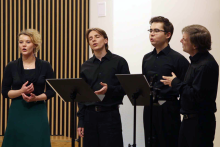 Cztery osoby ubrane na czarno stoją w półokręgu, śpiewają lub przemawiają. Przed nimi dwa pulpity na nuty lub teksty. W tle pionowe drewniane listwy po lewej i biała ściana po prawej stronie.