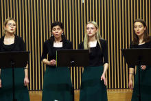 Cztery kobiety w ciemnozielonych spódnicach i czarnych bluzkach stoją na scenie, śpiewają lub przygotowują się do śpiewu. Przed nimi trzy statywy z nutami. W tle drewniana ściana z pionowymi pasami w ciemnych i jasnych odcieniach.