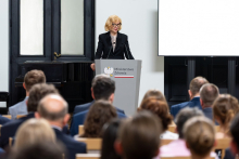 Kobieta, wiceminister zdrowia, przemawia za mównicą z logo Ministerstwa Zdrowia. Kobieta ma jasne włosy, okulary, jest elegancko ubrana w czarny garnitur. Przed nią siedzi grupa ludzi zwróconych w jej stronę. W tle widoczne są drzwi oraz ekran projekcyjny po prawej stronie.