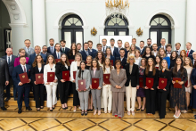 Około 40 osób w formalnych strojach. Uśmiechają się do zdjęcia. Stoją w eleganckim wnętrzu z jasnym parkietem, wysokim sufitem i ozdobnym żyrandolem. Większość trzyma czerwone teczki z godłem Polski. W tle widoczne są trzy duże, półokrągłe okna oraz białe kolumny.