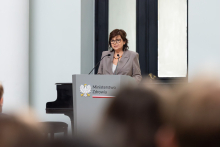 Kobieta, minister zdrowia, przemawia za mównicą z logo Ministerstwa Zdrowia. Kobieta ma ciemne włosy, okulary, jest elegancko ubrana w szary garnitur. W tle widoczne są fortepian i duże okno. 