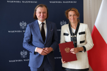 Dwie osoby stoją na tle granatowej ściany z napisem „WOJEWODA MAZOWIECKI” i godłem Polski. Osoba po lewej ma niebieski garnitur, białą koszulę i ciemny krawat. Osoba po prawej ma biały żakiet z czarnymi elementami i trzyma czerwoną teczkę z godłem. Po prawej stronie widoczna jest flaga Polski.