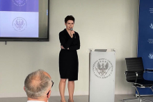  Kobieta, kanclerz WUM, w czarnej sukience przemawia do mikrofonu przy mównicy w sali konferencyjnej. W tle ekran z logo Warszawskiego Uniwersytetu Medycznego i krzesło biurowe. Na pierwszym planie widoczna głowa uczestnika siedzącego tyłem do aparatu
