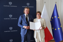 Dwie osoby stoją przed ciemnoniebieskim tłem z napisem „WOJEWODA MAZOWIECKI” i godłem Polski. Osoba po lewej ma niebieski garnitur, białą koszulę i krawat. Osoba po prawej ma jasny garnitur i trzyma otwartą teczkę z dokumentem. W tle widoczne są flagi Polski i Unii Europejskiej.