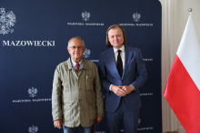 Dwóch mężczyzn stoi na tle granatowej ścianki z napisem „WOJEWODA MAZOWIECKI” i godłem Polski. Mężczyzna po lewej ma zieloną kurtkę i czerwony krawat. Mężczyzna po prawej ma niebieski garnitur, białą koszulę i ciemny krawat. Po prawej stronie widoczna jest flaga Polski.