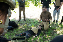 A group of people outdoors during training. In the foreground, a person wearing a protective vest is lying on the grass, while another kneels next to him, performing a rescue action. In the background, several people in tactical gear are observing the situation. The scene takes place under a tree.
