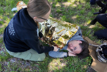 Students during a training ground exercise. A girl in a black sweatshirt kneels on the grass and covers a person lying down with a golden emergency blanket. Other people's legs and a backpack on the ground are visible in the background.