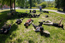 Students during exercises at the training ground. A group of people lie on the grass in the park. A path and a brick building are visible in the background. The individuals are wearing dark clothes, with backpacks lying next to them. 