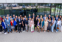 Grupa około 30 osób stoi przed budynkiem z dużymi, przeszklonymi oknami. Ustawieni w dwóch rzędach, patrzą w stronę aparatu. W tle odbicia ludzi, drzewa i zaparkowane samochody. Twarze zamazane, stroje eleganckie i casualowe.