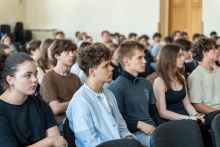 Grupa młodych osób siedzących w sali wykładowej, zwróconych twarzami w stronę prelegenta lub prezentacji poza kadrem. W tle widoczne są rzędy krzeseł oraz jasne ściany.