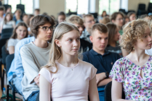 Grupa licealistów siedzących w sali wykładowej, skupionych na wykładzie lub prezentacji poza kadrem. W tle widoczne są kolejne rzędy uczestników oraz okna wpuszczające naturalne światło.