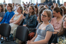 Grupa licealistów siedzących w sali wykładowej, zwróconych w stronę mównicy, uczestniczących w wykładzie. W tle widoczne są inne osoby oraz okna wpuszczające naturalne światło.