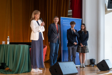 Cztery osoby stojące na scenie na tle niebieskiej ścianki z napisem „MEDICAL UNIVERSITY OF WARSAW”. Po lewej kobieta w białej marynarce i niebieskiej sukience przemawia do mikrofonu. Obok niej mężczyzna w ciemnej marynarce, czerwonej koszuli i dżinsach. Po prawej stronie chłopak w ciemnym garniturze oraz dziewczyna w mundurku szkolnym z ciemną marynarką i spódnicą w kratę. 