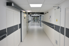 Jasny szpitalny korytarz z białymi ścianami i szarymi pasami. Na końcu szyba. Po lewej stronie drzwi z napisem „PRACOWNIA HEMODYNAMIKI I”. Na suficie prostokątne lampy.