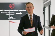 Mężczyzna w garniturze, prof. Marcin Grabowski, trzyma kartkę papieru. Obok niego częściowo widoczna druga osoba. W tle czarny szyld z białym napisem: „Oddział i Pracownie Kardiologii Inwazyjnej, Pracownie Zabiegowe i Sale Chorych”. Na szyldzie czerwone serce z linią EKG i strzałka wskazująca kierunek.