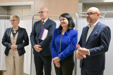 Cztery osoby stoją obok siebie na tle białych parawanów. Pierwsza osoba w jasnym garniturze i ciemnej marynarce. Druga w ciemnym garniturze, trzyma dokumenty. Trzecia w niebieskiej marynarce. Czwarta w ciemnym garniturze, trzyma zwinięty dokument.