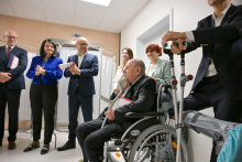 Grupa osób w eleganckich strojach stoi w jasnym pomieszczeniu z białymi ścianami. W tle białe parawany medyczne. Na pierwszym planie mężczyzna na wózku inwalidzkim trzyma kule. Obok niego stoi inny mężczyzna, opierając się na kulach.