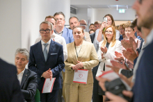 Grupa elegancko ubranych osób stoi w jasnym korytarzu. Niektórzy trzymają dokumenty z czerwonym sercem na okładce. Twarze zamazane. W tle drzwi ewakuacyjne z zielonym znakiem wyjścia. Część osób klaszcze.