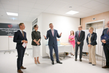 Sześć osób stoi w jasnym pomieszczeniu. Trzy po lewej ubrane formalnie, dwie trzymają dokumenty. Na środku mężczyzna w garniturze gestykuluje. Po prawej trzy osoby, jedna z teczką. W tle stół z napojami i przekąskami oraz tablica informacyjna o oddziale kardiologii inwazyjnej.