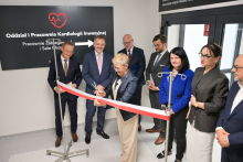 Grupa elegancko ubranych osób stoi w jasnym pomieszczeniu. Kobieta w środku- prof. Maria Mrówczyńska, podsekretarz stanu w Ministerstwie Nauki i Szkolnictwa Wyższego - przecina biało-czerwoną wstęgę nożyczkami. W tle czarna tablica z napisem „Oddział i Pracownia Kardiologii Inwazyjnej”, czerwoną grafiką serca z EKG i strzałką kierunkową.