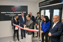 Grupa osób stoi przed wejściem do Oddziału Kardiologii Inwazyjnej. Kobieta w szarym żakiecie - Marzena Kowalczyk, dyrektor UCK WUM - przecina czerwoną wstęgę, trzymaną przez dwóch mężczyzn. Po lewej czarna tablica z białym napisem o pracowniach i salach chorych. Po prawej drzwi z podobnym napisem.