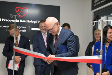 Mężczyzna - prof. Rafał Krenke, rektor WUM - w garniturze przecina czerwoną wstęgę. Obok stoją formalnie ubrane osoby. W tle czarna tablica z białym napisem „Pracownia Kardiologii” i czerwonym sercem z linią EKG. Po prawej stronie ściany widoczny napis „ODDZIAŁ PRACOWNIE HEMODYNAMIKI”. Pomieszczenie jest jasne.