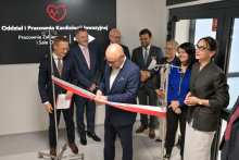 Mężczyzna - Filip Nowak, prezes Narodowego Funduszu Zdrowia - w garniturze przecina biało-czerwoną wstęgę podczas otwarcia oddziału. W tle czarna tablica z białym napisem: „Oddział i Pracownia Kardiologii Inwazyjnej, Pracownie Zabiegowe i Sale Hybrydowe”. Obok elegancko ubrani uczestnicy, część trzyma dokumenty.