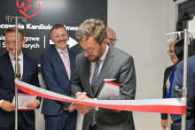 Mężczyzna - prof. Paweł Włodarski, dziekan Wydziału Lekarskiego - w garniturze przecina biało-czerwoną wstęgę nożyczkami. W tle osoby w formalnych strojach. Za nimi czarny baner z białym napisem „Pracownia Kardiologii Inwazyjnej” i czerwonym sercem z linią EKG.