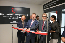 Trzech mężczyzn w garniturach - prof. Grabowski, prof. Kochman i prof. Opolski - przecina czerwoną wstęgę. W tle czarna tablica z białym napisem „Oddział i Pracownia Kardiologii Inwazyjnej, Pracownie Zabiegowe i Sale Chorych” oraz czerwonym sercem z linią EKG. Po prawej wejście z napisem „Oddział Kliniczny Kardiologii, Pracownie Hemodynamiki, Sale Chorych”.