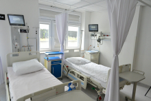 Jasny pokój szpitalny z dwoma białymi łóżkami, poduszkami i pościelą. Nad łóżkami monitory medyczne. Pomiędzy łóżkami niebieski stolik na kółkach. W tle dwa duże okna z białymi roletami i zasłona na szynie. Po prawej stronie ściany sprzęt medyczny i mała szafka.