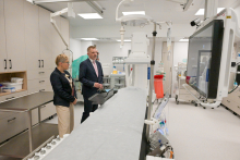 Dwie formalnie ubrane osoby stoją w nowoczesnym gabinecie medycznym. Jedna trzyma dokumenty. Po prawej stronie duży monitor, po lewej sprzęt medyczny i szafki. Dominują kolory biały i szary.