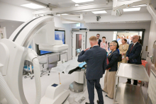 Nowoczesna sala operacyjna z białymi ścianami i sufitem. Na środku stół operacyjny przykryty niebieskim materiałem. Z przodu duży aparat z ramieniem w kształcie litery C. Kilka osób w garniturach ogląda sprzęt. Jedna osoba obsługuje panel dotykowy. Jasne oświetlenie sufitowe.