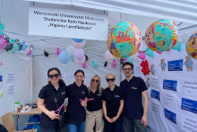 Promotional tent of the Warsaw University of Medical Sciences, Students' Scientific Circle “Hygiene and Prevention”. Inside, five people wearing navy blue T-shirts with the WUM logo. Above them are colourful balloons, including two with characters from the ‘Paw Patrol’ cartoon. Information posters about hand hygiene are visible on the walls of the tent.