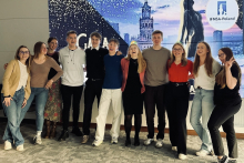 Dziesięć osób stoi w rzędzie na tle dużego ekranu z grafiką miasta i logo IFMSA-Poland. Uczestnicy mają stroje od codziennych po formalne. Podłoga pokryta jest szarą wykładziną.