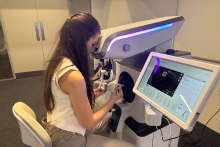 Młoda kobieta siedzi na krześle i obsługuje białe urządzenie medyczne z niebieskim podświetleniem. Patrzy przez okular mikroskopu i trzyma ręce na kontrolerach. Przed nią znajduje się duży ekran dotykowy z danymi i obrazami. W tle widoczne są drzwi i fragment biura.