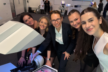 Grupa pięciu kobiet w formalnych ubraniach stoi wokół dużego, białego urządzenia medycznego skierowanego w dół. Patrzą na ekran z obrazem medycznym. W tle widać salę konferencyjną, inne osoby i sprzęt elektroniczny.