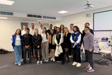 Piętnaście osób stoi w jasnej sali konferencyjnej. W tle widać biały ekran projekcyjny, krzesła i sprzęt elektroniczny. Część osób ma formalne ubrania, inni są ubrani swobodnie. Ściany są białe, a podłoga pokryta szarym dywanem.