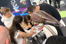 Grupa osób stoi wokół stołu z urządzeniem medycznym. Osoby patrzą na sprzęt i wskazują go palcami. Na pierwszym planie kobieta w jasnej bluzce, ciemnej kamizelce i chustce na głowie. W tle plakat z grafiką sprzętu medycznego i kolorowymi elementami. Jedna osoba ma zasłoniętą twarz.