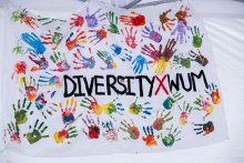 Biały baner pokryty kolorowymi odciskami dłoni w barwach czerwieni, niebieskiego, zieleni, żółci i fioletu. W centrum widoczny napis „DIVERSITY X WUM”, gdzie litera „X” jest czerwona, a pozostałe czarne.