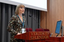 Kobieta w sukience we wzory stoi za mównicą z napisem „VERITATIS SPLENDOR”. Na szyi ma identyfikator, przed nią ustawione są dwa mikrofony. W tle widoczne są zasłony i fragment ekranu projekcyjnego. Po prawej stronie znajduje się monitor z logo „SAPIENTIAE STUDIUM”.