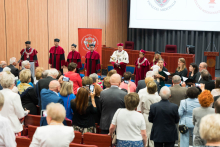 Uroczystość w auli. Na scenie siedem osób: władze rektorskie i dziekańskie w togach i biretach, jeden trzyma otwartą księgę. W tle czerwony baner z godłem wydziału. Na pierwszym planie uczestnicy robią zdjęcia władzom.