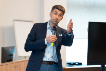 A man in a navy blue jacket and light blue shirt holds a microphone. He stands in a conference room next to a white board and monitor. He speaks, gesturing with his right hand.