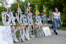 Duże litery tworzącymi napis Diversity x WUM, ozdobione mozaikowymi, kwiatowymi wzorami. W tle dwie osoby trzymają papierowe kwiaty. Po prawej kobieta w białej bluzce i niebieskiej spódnicy. W tle drzewa i fragmenty budynków.