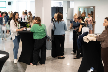 Grupa osób rozmawia przy wysokich stolikach koktajlowych w jasnej, przestronnej sali konferencyjnej. W tle widoczne są inne osoby oraz stoiska z materiałami. Na stolikach znajdują się m.in. rośliny doniczkowe i torby na zakupy. Przestrzeń oświetlona jest naturalnym światłem z dużych okien.