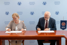Po lewej kobieta, dyrektor NIO, po prawej mężczyzna rektor WUM, siedzą przy drewnianym stole i podpisują dokumenty. W tle niebieska ściana z logo i napisem „Warszawski Uniwersytet Medyczny”. Kobieta ma na sobie jasny sweter, mężczyzna ciemny garnitur z bordowym krawatem. Na stole widoczna flaga uczelni.