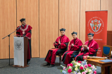 Mężczyzna w czerwono-czarnej todze - dziekan WL przemawia przy mównicy z logo uczelni. Obok siedzą trzy osoby w togach, klaszczące. W tle czerwony baner z logo Wydziału Lekarskiego. Na pierwszym planie stół z bukietem kwiatów.